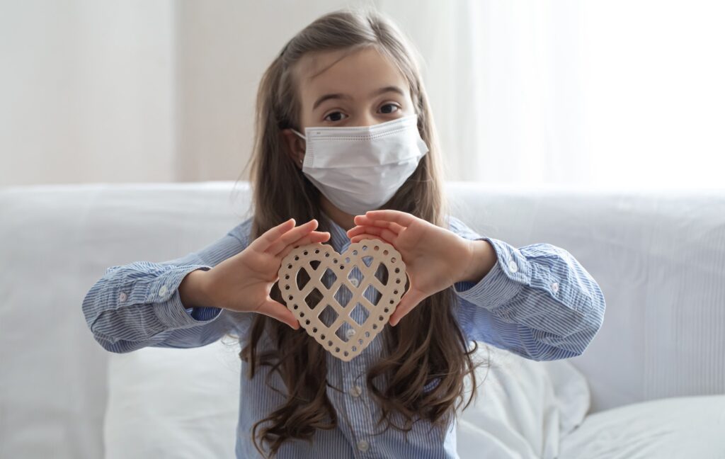 Little girl in a protective mask with a wooden decorative heart in her hands.